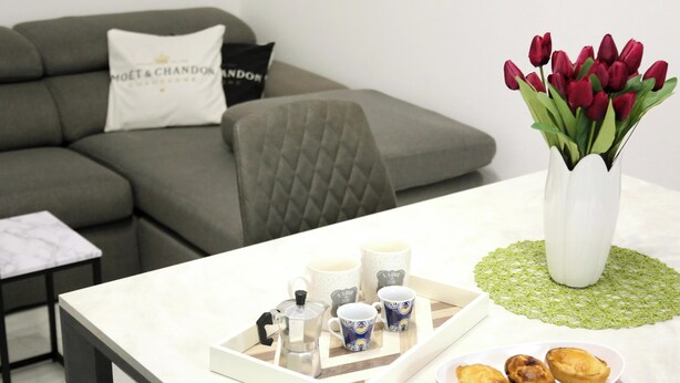 Ein modernes Wohnzimmer mit einem grauen Sofa, einem weißen Couchtisch, einer Auflage mit einer Kaffeemaschine, Tassen und Gebäck, sowie einer Vase mit roten Tulpen.
