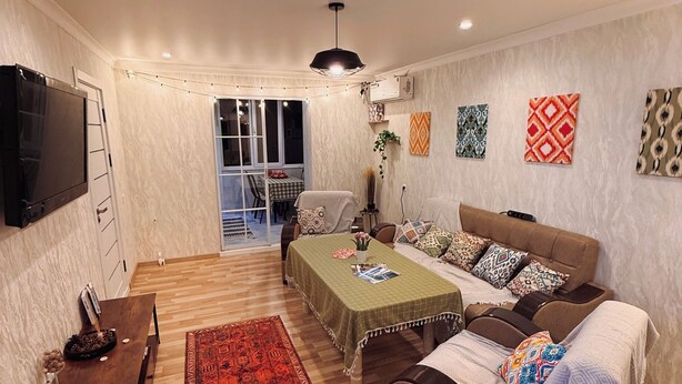 Una sala de estar con un sofá, una mesa de centro, un televisor, un comedor con mesa y sillas, y un tapete rojo estampado.