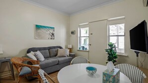 A living room with a grey sofa, wicker chairs, a round table with a bowl, a painting on the wall, and a TV on the wall. - No 11 The Smugglers Inn (Victor Harbor)