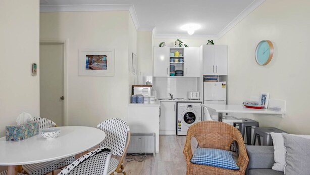 A compact kitchen with white cabinets, a built-in washing machine, and a dining area with a round table and patterned chairs. - No 11 The Smugglers Inn (Victor Harbor)
