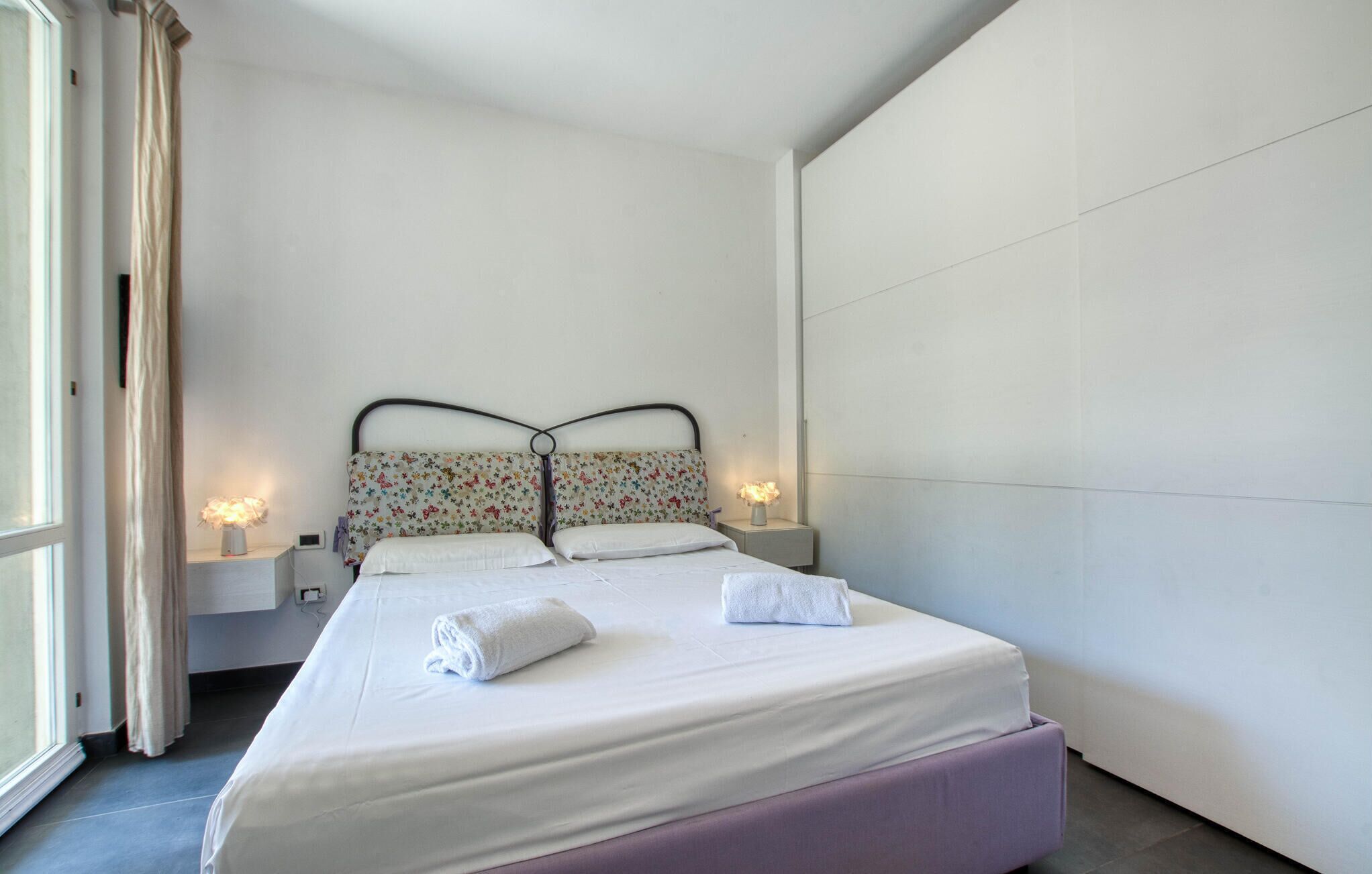 Un dormitorio con una cama, dos lámparas de noche, una ventana con cortinas y una mesita de noche.