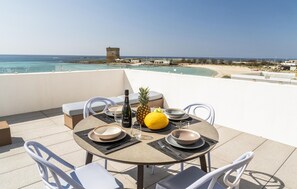 Ein runder Tisch mit vier Stühlen, eine Weinflasche, eine Ananas, eine Zitrone und eine Schüssel auf einer Terrasse mit Blick auf einen Strand und das Meer.