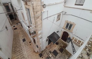 Ein schmaler Innenhof mit Steinboden, weißen Wänden und einem kleinen Balkon mit Metallgeländern.
