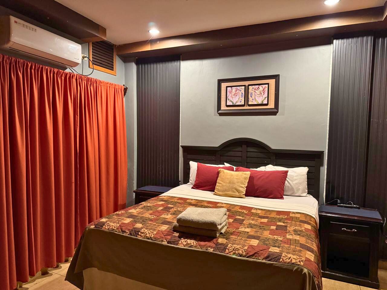 A hotel room with a bed, a nightstand, a window, and a wall-mounted air conditioner.