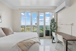 A bedroom with a bed, a desk, and chairs, a large window with a view, and an air conditioning unit. - Condado Lagoon - Lux Waterfront Studios (San Juan)