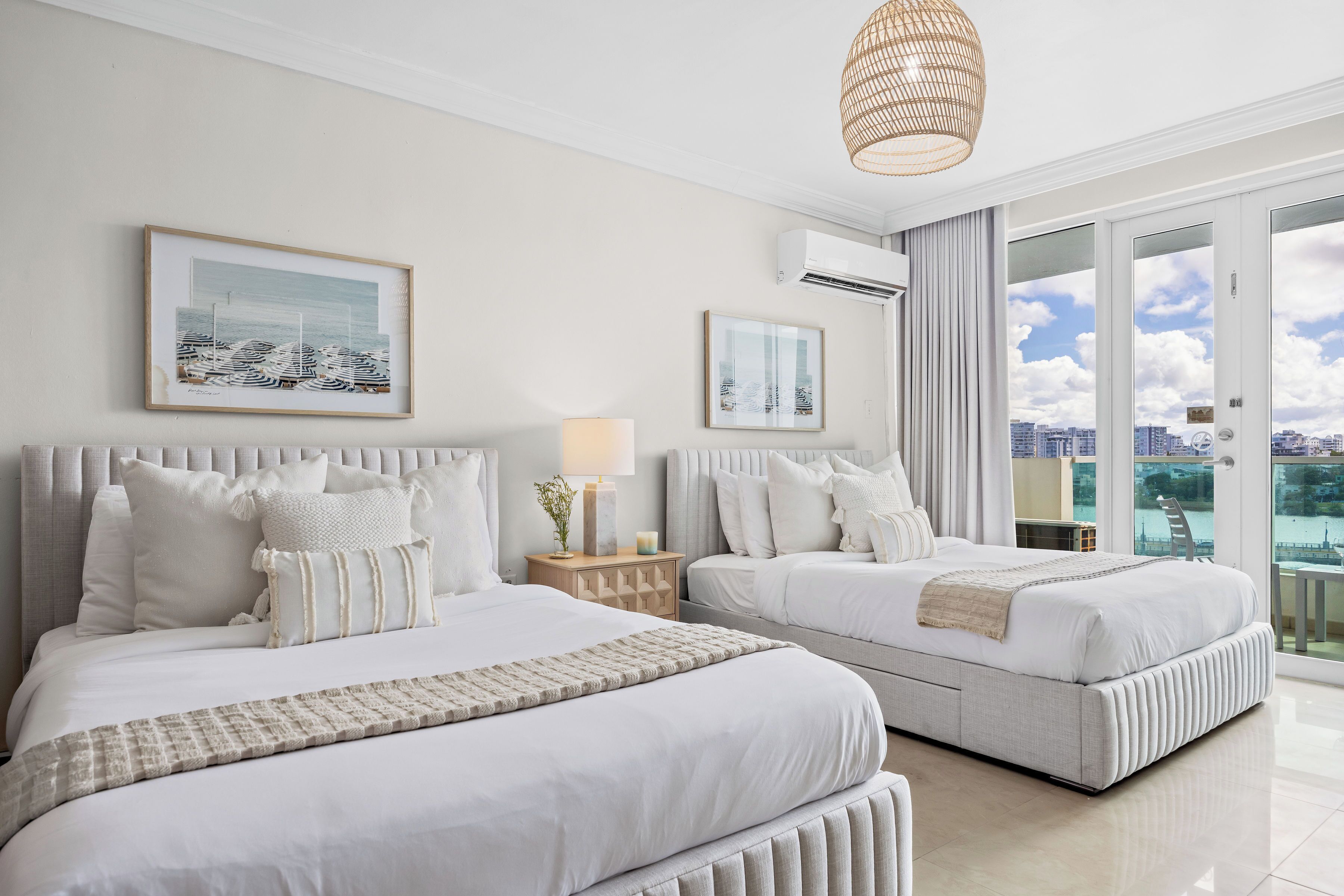 Quarto de hotel com duas camas, uma janela grande com vista para a cidade, um ventilador de teto e obras de arte nas paredes.