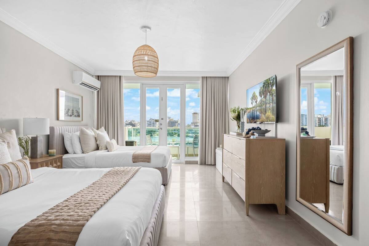 Habitación de hotel con dos camas, un espejo grande, un aparador y vista a la ciudad.