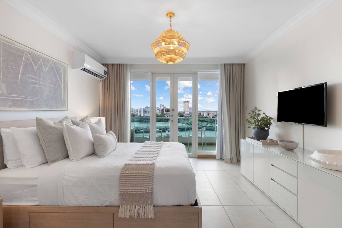 Un dormitorio moderno con una cama grande, televisión y vistas de la ciudad a través de las puertas del balcón.