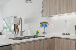 A modern kitchen with a double sink, wooden cabinets, a stainless steel faucet, a small plant, and a box of chips on the counter. - Stylish Pool Home Near Disney | BBQ + Spacious Retreat for 10 (Clermont)