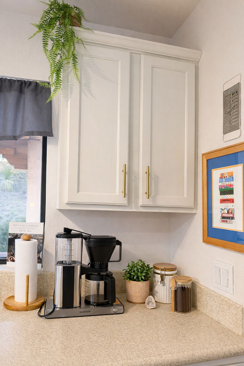 Een keuken met witte kastjes, een koffiezetapparaat, een bloempot en een ingelijst schilderij aan de muur.