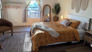 Ein Schlafzimmer mit Bett, Stuhl, Deckenventilator, Fenster mit Vorhängen, Spiegel, Pflanze und Wandbehang aus Makramee.