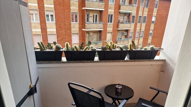 Een balkon met een kleine tafel en stoelen, potten met cactussen en uitzicht op een bakstenen gebouw met meerdere balkons.