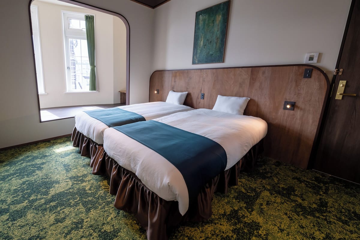 A hotel room with two beds, a window with curtains, a wooden headboard, and a green carpet.