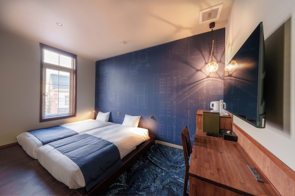 A bedroom with a bed, a desk, a chair, a window, and a wall with architectural drawings.