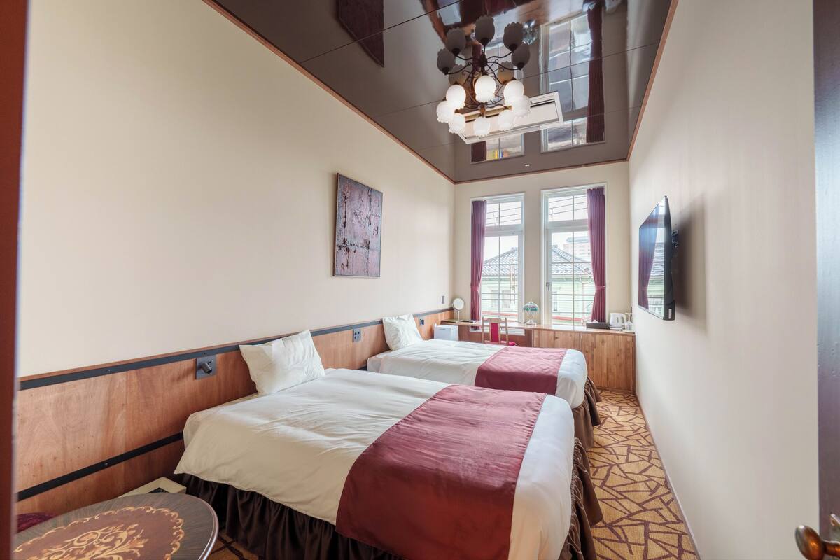 A hotel room with two beds, a ceiling fan, a flat-screen TV, and a window with curtains.
