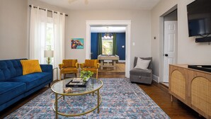Ein Wohnzimmer mit einem blauen Sofa, einem runden Couchtisch, einem Essbereich mit Tisch und Stühlen sowie einem an der Wand montierten Fernseher.