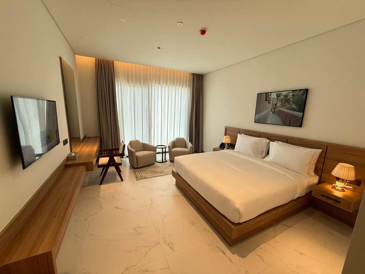 Ein Hotelzimmer mit einem großen Bett, einem Fernseher, einem Schreibtisch, einem Stuhl, einer Lampe und einem Bild an der Wand.