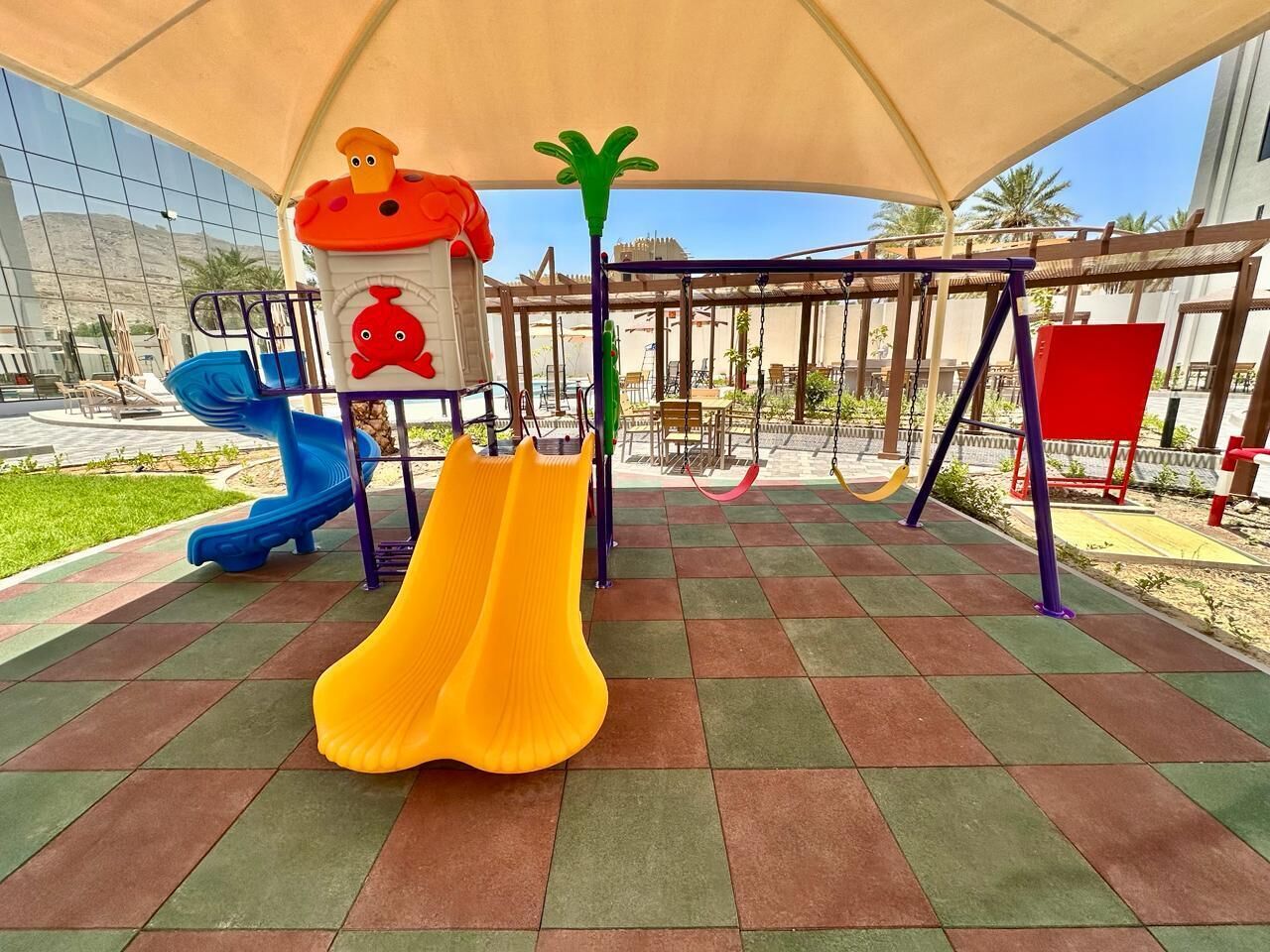 Un terrain de jeux avec des toboggans et des balançoires, installé sous une sorte de tonnelle ou de pergola.