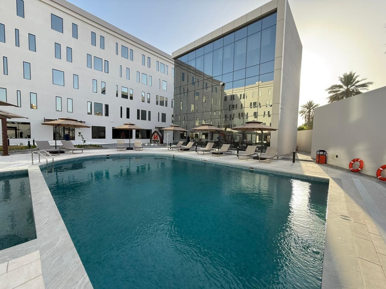A swimming pool with lounge chairs and umbrellas, surrounded by a hotel building with multiple floors and large windows.