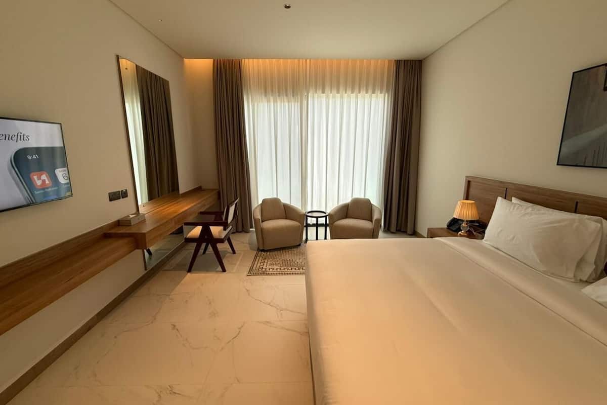 A hotel room with a large bed, a desk, two armchairs, a small table, a TV, and a window with curtains.