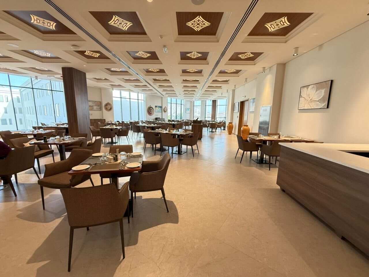 A spacious hotel dining area with multiple tables and chairs, a reception desk, and large windows providing natural light.