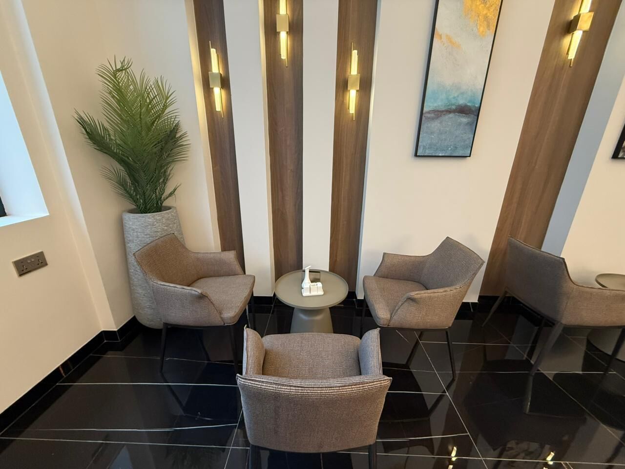 A lobby with a black-tiled floor, three grey armchairs, a small round table, a potted plant, vertical light fixtures, and a painting on the wall.