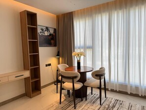 A modern room with a round dining table, two chairs, a wooden bookshelf, a painting on the wall, and a large window with curtains. - Swiss-Belhotel Nizwa (Nizwa)