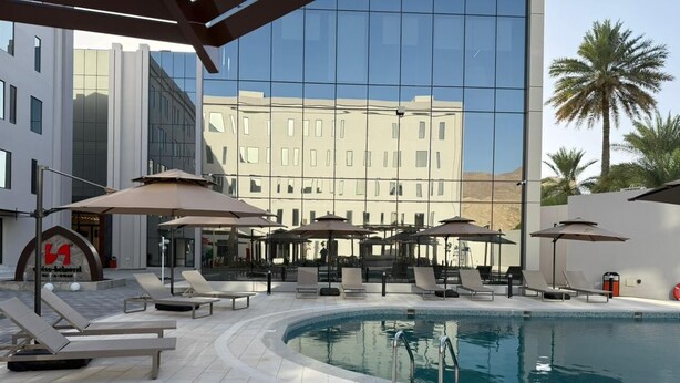 Zona de piscina con sillones de descanso y sombrillas, un edificio moderno con ventanas reflectantes y palmeras al fondo.
