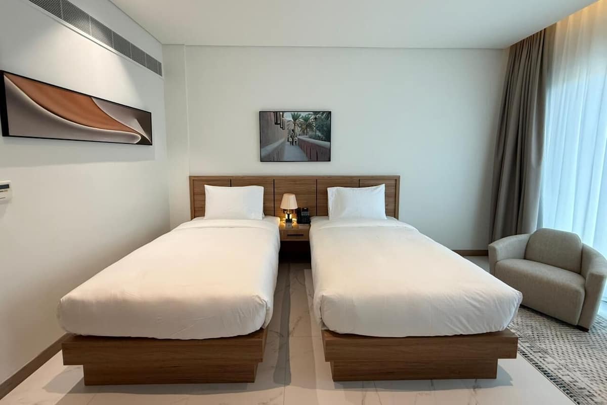 A hotel room with two single beds, a nightstand with a lamp, a chair, and a painting on the wall.