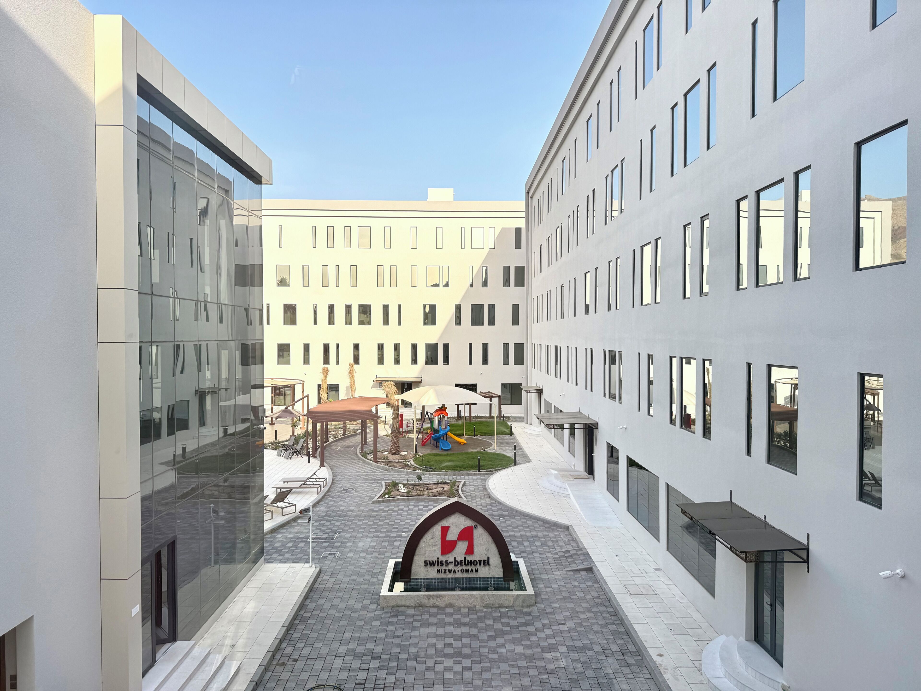 A modern hotel courtyard with a central sign, outdoor seating, and a children's play area.