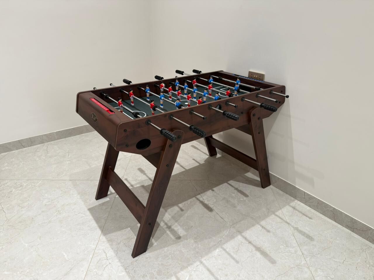 Una mesa de fútbol de madera con estructura metálica, frente a una pared lisa con piso de baldosas.