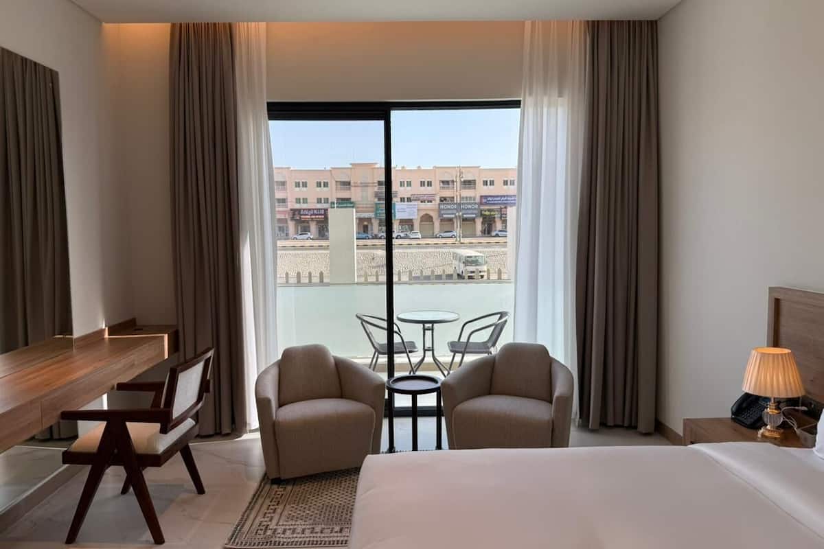 A hotel room with a bed, two armchairs, a small table, a desk, and a view of a cityscape through large windows.