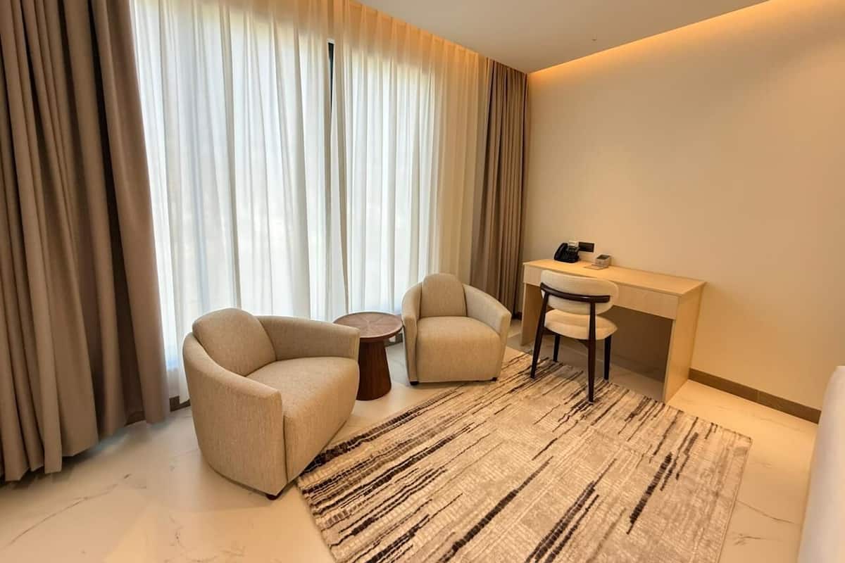 A hotel room with two armchairs, a small round table, a desk with a chair, and a large window with curtains.