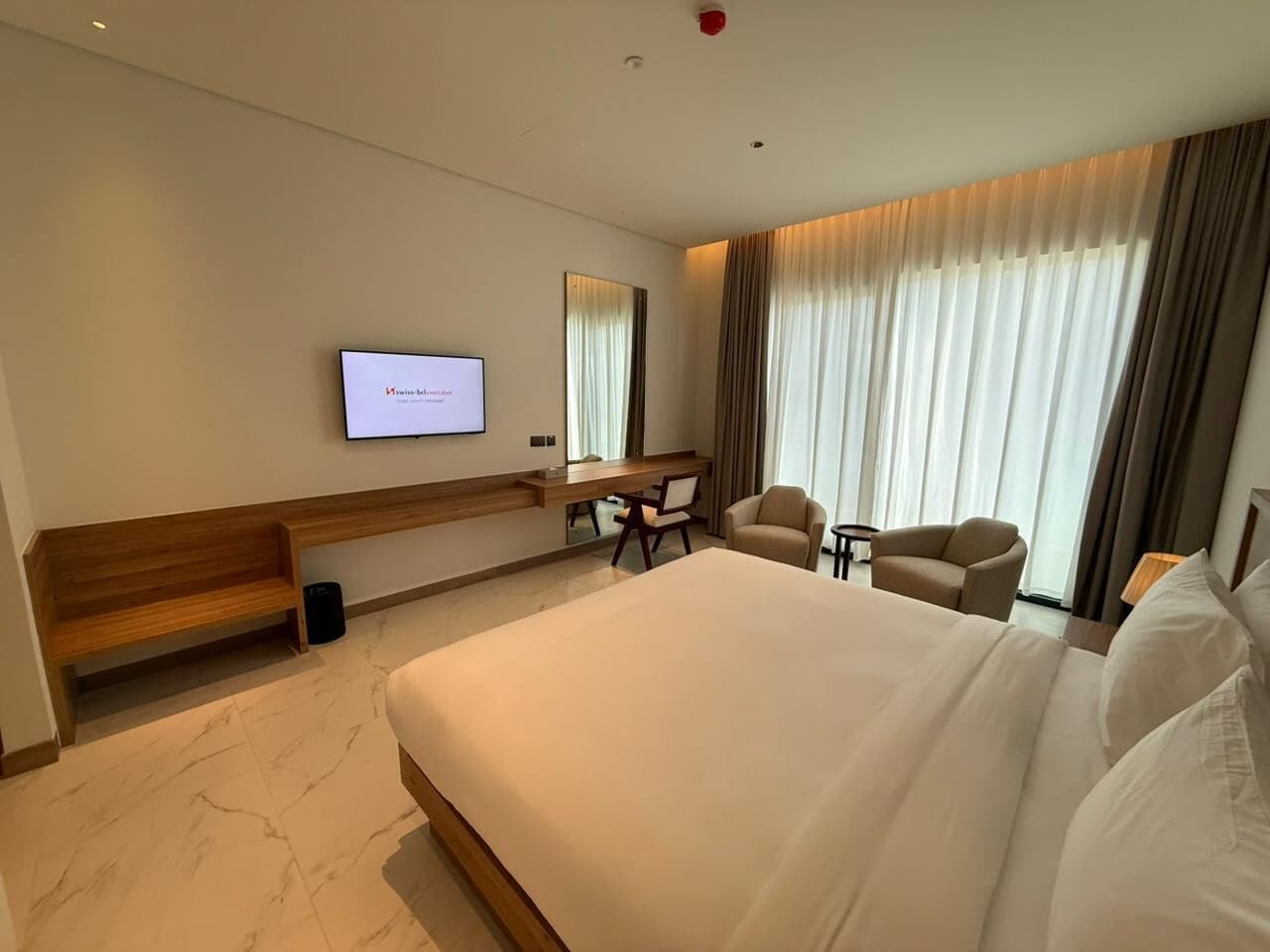 A hotel room with a large bed, a desk with a chair, a television, and a view of the outdoors through curtains.