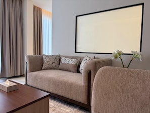 Une chambre d’hôtel moderne avec un canapé beige, une table basse en bois, une grande œuvre d’art encadrée au mur et un vase rempli de fleurs.