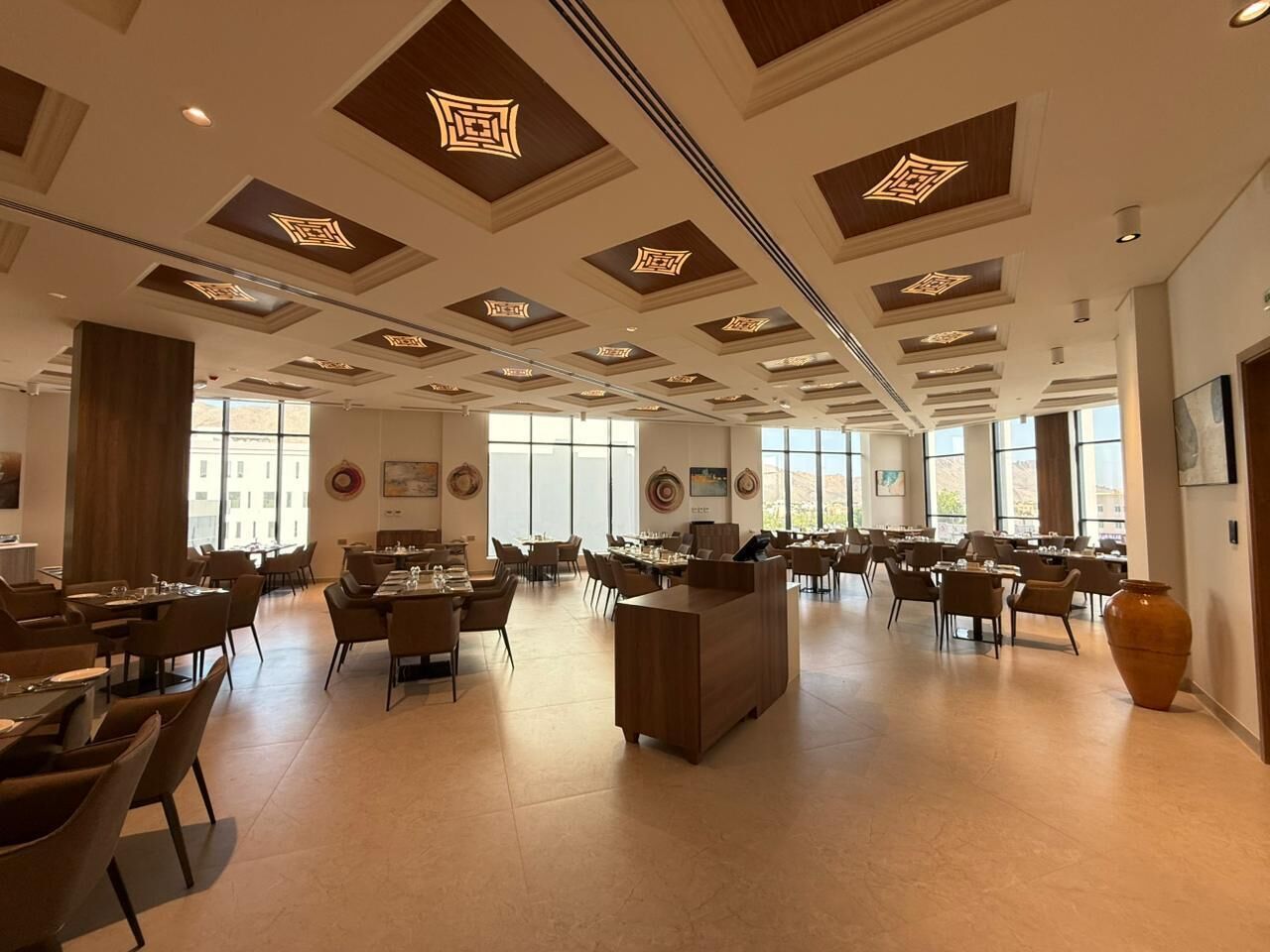A spacious hotel lobby with multiple dining areas, a reception desk, and large windows allowing natural light.