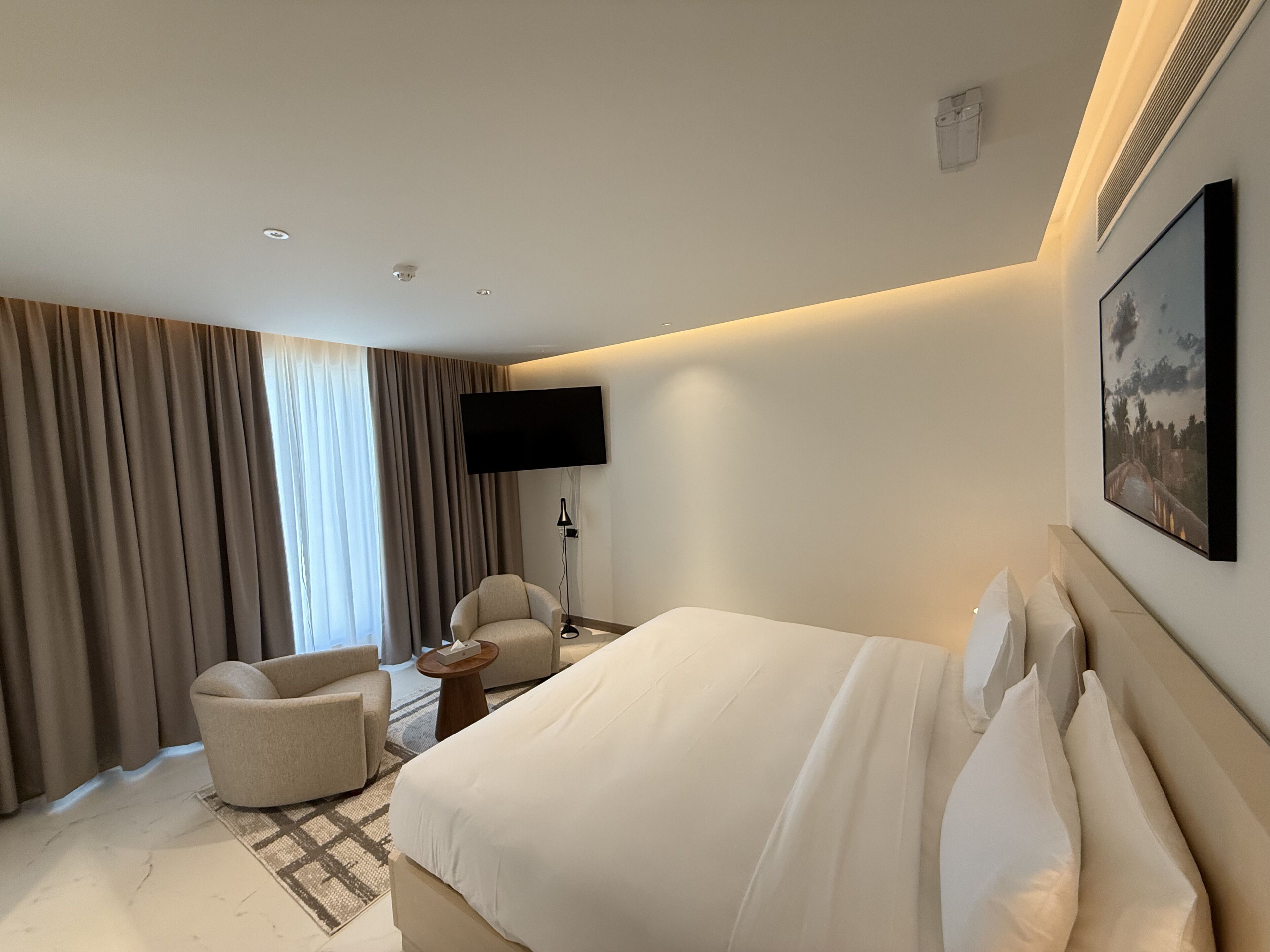 A hotel room with a bed, a chair, a TV, and a painting on the wall.