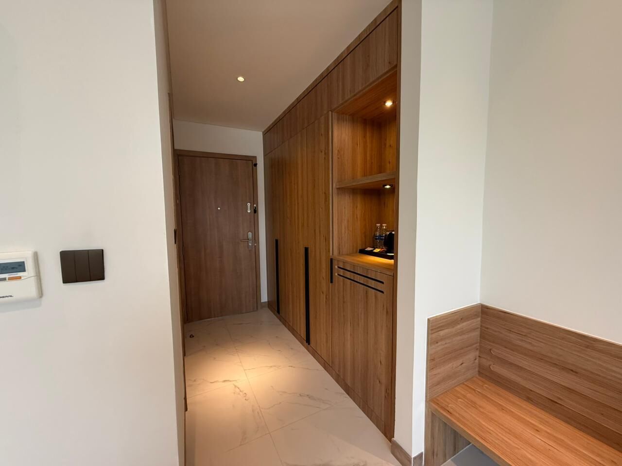 Un couloir avec une armoire en bois, une petite étagère sur laquelle sont posés quelques objets, et un thermostat fixé au mur.