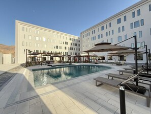 Un espace piscine avec des chaises longues et des parasols, un ciel dégagé et un bâtiment moderne doté de nombreuses fenêtres.