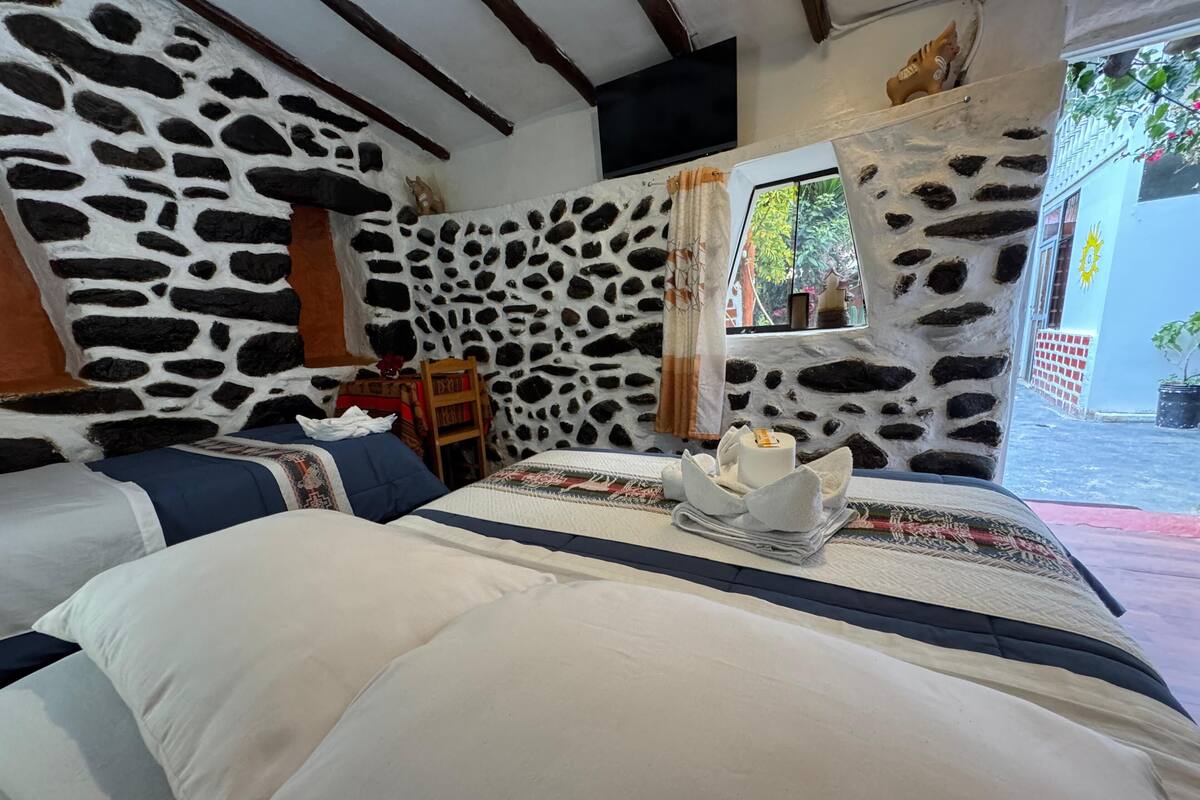 Un dormitori amb paret de pedra, llit amb roba de llit blanca, taula petita amb una làmpada i finestra amb cortines.