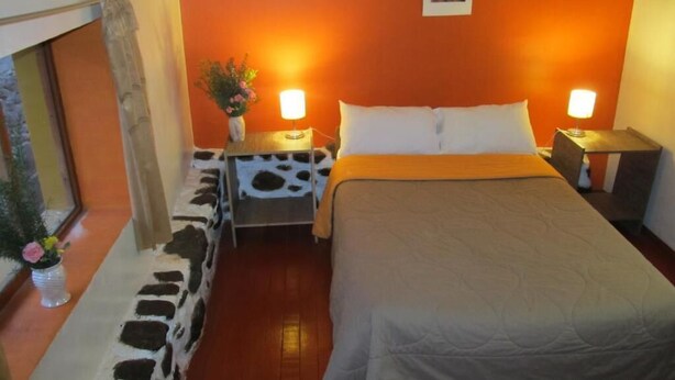 A room with a bed, two bedside tables, a nightstand, a window with a curtain, and a wall-mounted picture. - Killari B&B (Ollantaytambo)