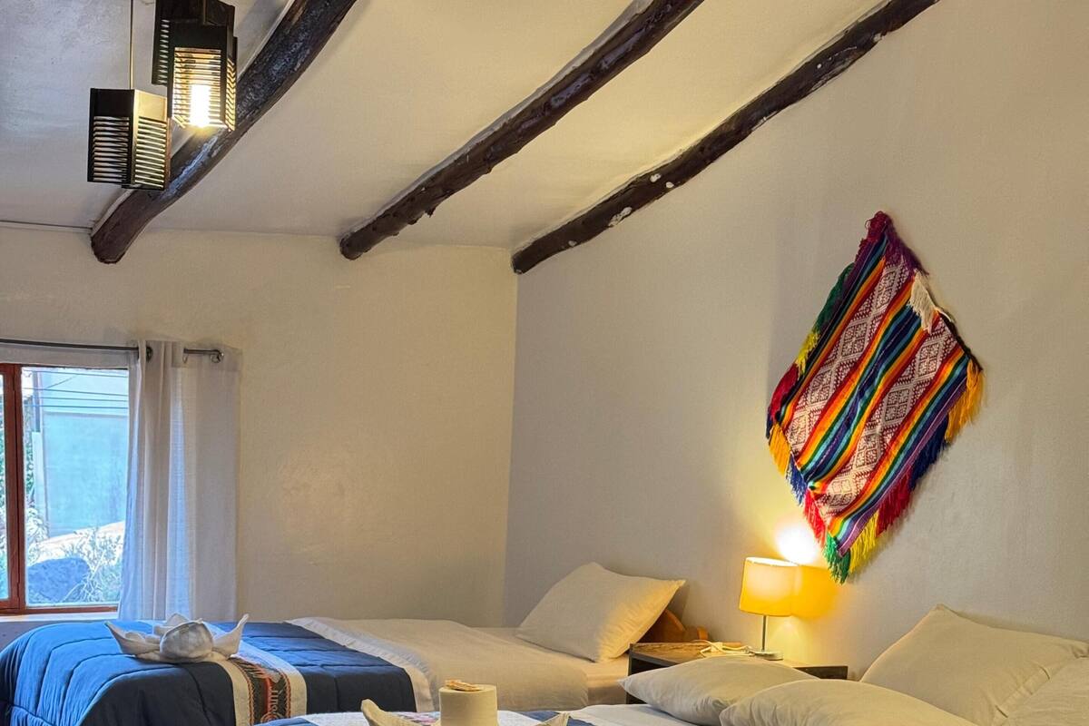 Habitació d'hotel amb tres llits, una finestra amb cortines, una manta colorida a la paret i una làmpada al taulell.