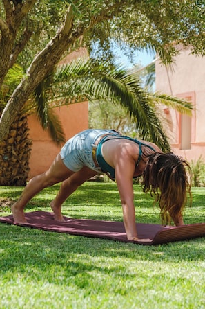 Una persona realizando una postura de yoga sobre una esterilla al aire libre, con árboles y un edificio de fondo.