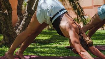 Una persona realizando una postura de yoga sobre una esterilla al aire libre, con árboles y un edificio al fondo.
