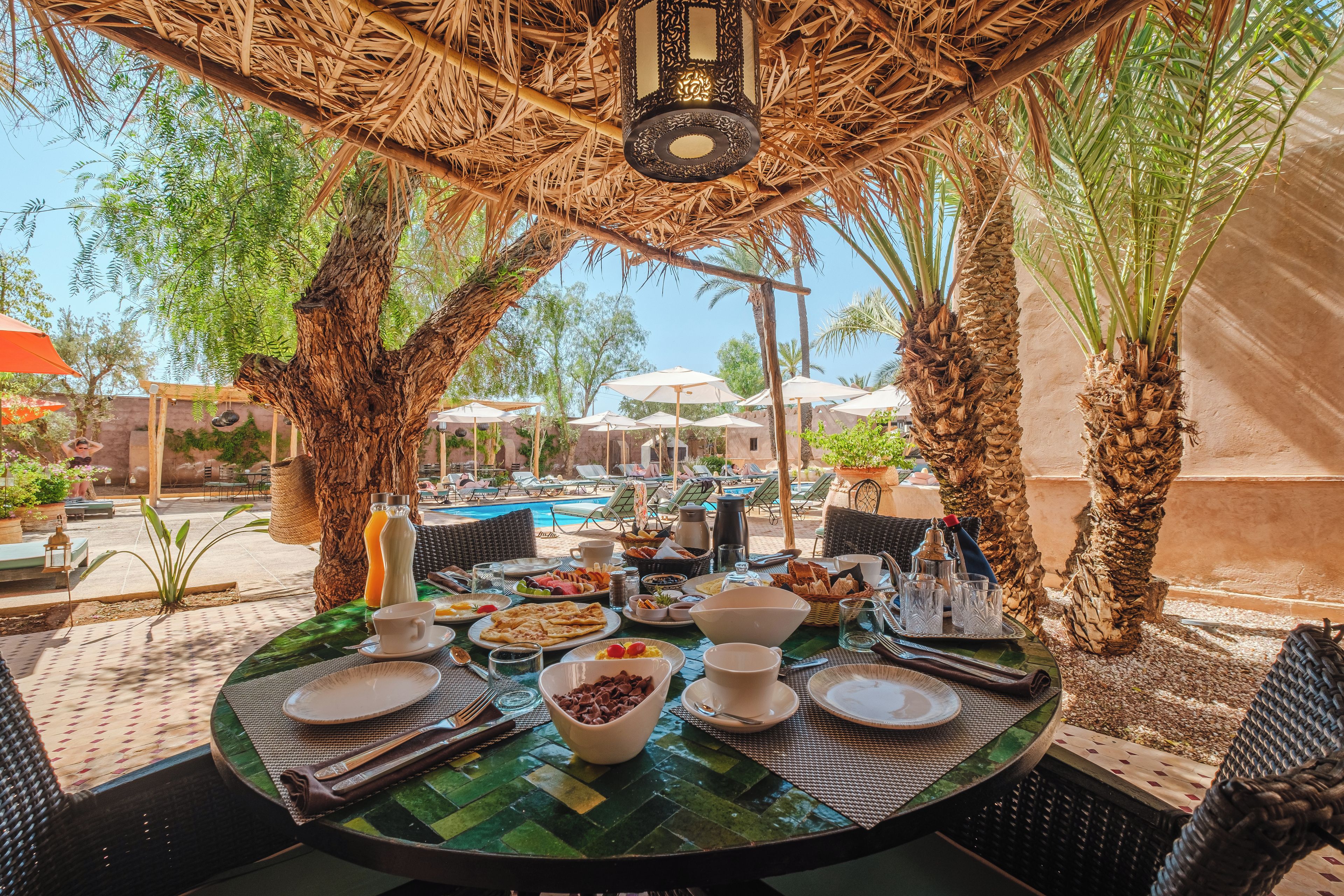 Un área de comedor al aire libre con vista a la piscina, rodeada de palmeras y sombrillas.