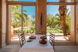A dining table with a white tablecloth, plates, glasses, and a bottle, set against a backdrop of palm trees and a clear sky. - Hotel Dar 55 Berbere House & Spa (Marrakech)