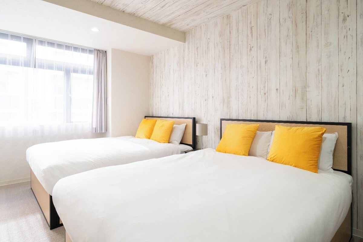 A hotel room with two beds, each with white bedding and yellow pillows, a wooden wall panel, and a window with curtains.