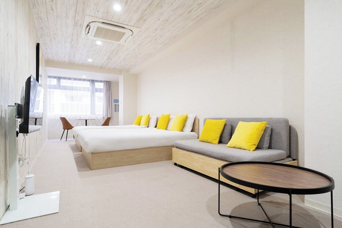 A modern hotel room with a large bed, a sofa with yellow cushions, a small round table, and a television.