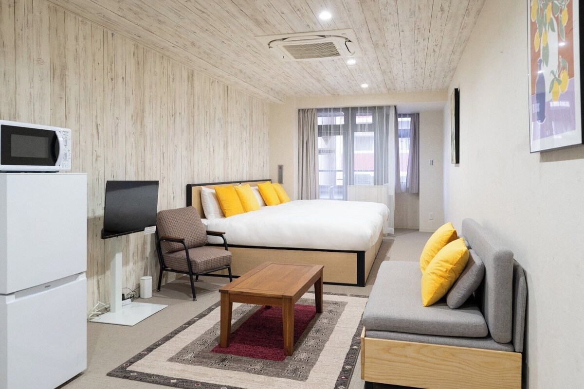 A modern hotel room with a bed, a sofa, a chair, a TV, and a refrigerator.
