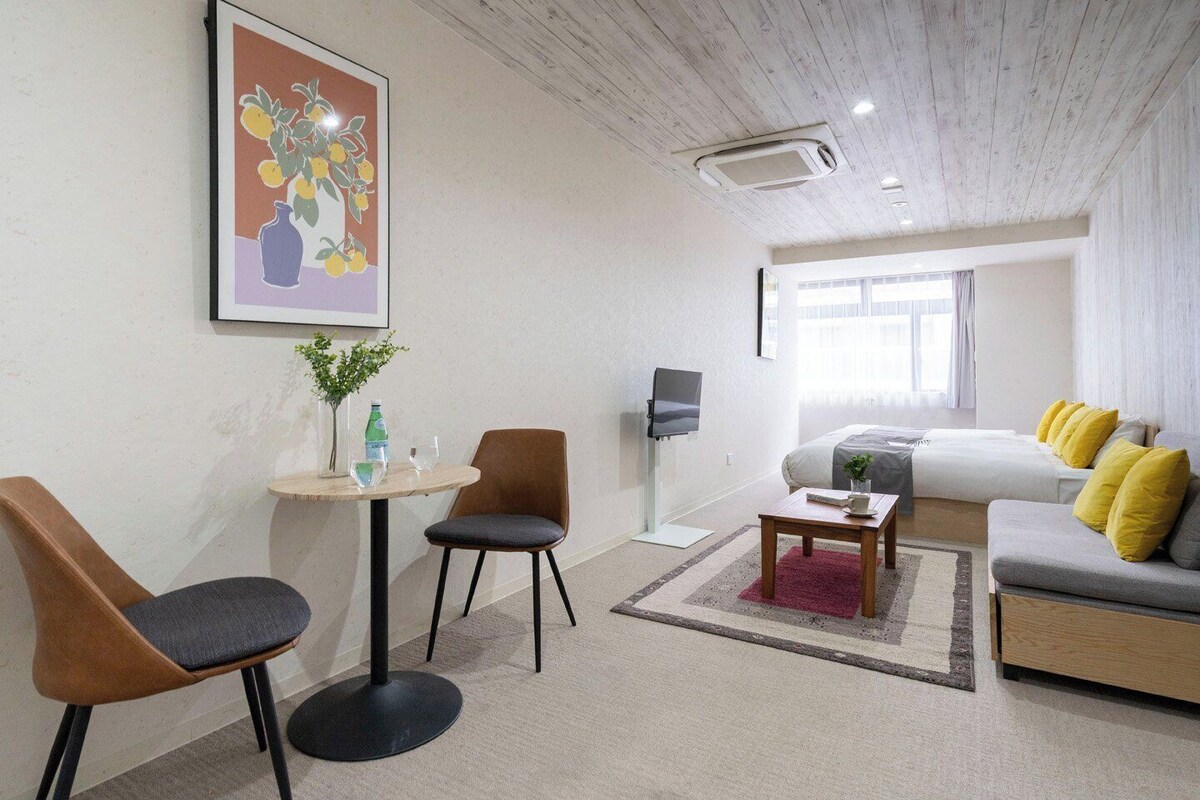 A modern hotel room with a bed, a sofa, a TV, a small table with a plant, and a framed picture on the wall.